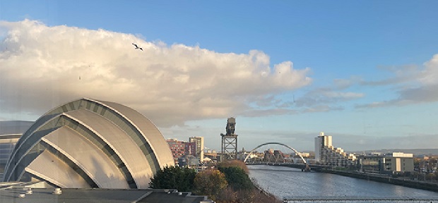 A photo of the ‘Armadillo’ conference centre, with the river Clyde, the Clyde Arc bridge and the city of Glasgow in the background, to the right. The sky is blue and a bird flies in front of a large, white, fluffy cloud.