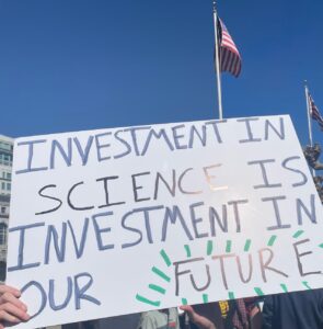 A white protest placard against a clear blue sky. Text on the placard reads, "Investment in science is investment in our future." The word 'future' is emphasised.