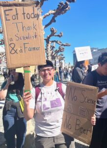 A person at a protest holds two cardboard placards, facing the camera. The text on one reads, "Eat today? Thank a scientist and a farmer." The second one reads, "No science, no beer."