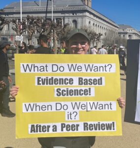 A person at a protest holds a yellow placard, facing the camera. The text on it reads, "What do we want? Evidence based science! When do we want it? After a peer review!"