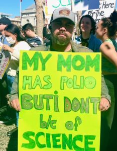 A person at a protest holds a yellow placard, facing the camera. The text on it reads, "My mom has polio but I don't, because of science."