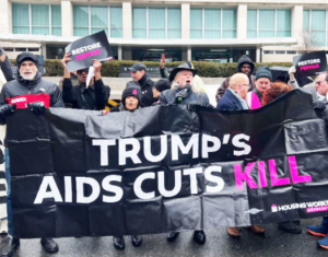 A group of people at a protest hold up a large black banner which has white and pink text on it. The text reads: Trump's AIDS cuts kill.