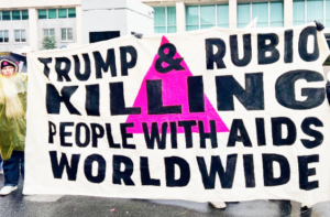 A group of people hold up a large white banner which has a pink triangle on it. Black text across it, reads: Trump and Rubio killing people with AIDS worldwide.