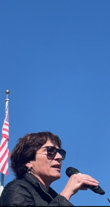 A white woman with short dark hair speaks into a microphone, outside. She is wearing sunglasses. There is a US flag behind her and the sky is bright and clear blue.