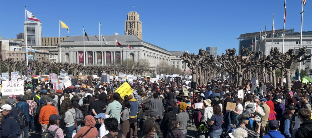 Stand Up For Science protest in San Francisco, 2025. A large crowd of people are gathered in front of Civic Centre Plaza on a sunny day. Some are holding placards. One says, "Stop Musk and the DOGE bros!"