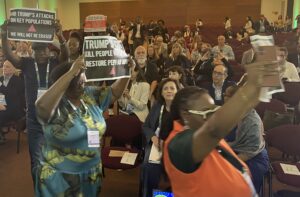 An action shot of protesters in front of delegates at the AIDS 2026 conference. The protesters are holding signs condemning Trump for PEPFAR and HIV funding cuts.